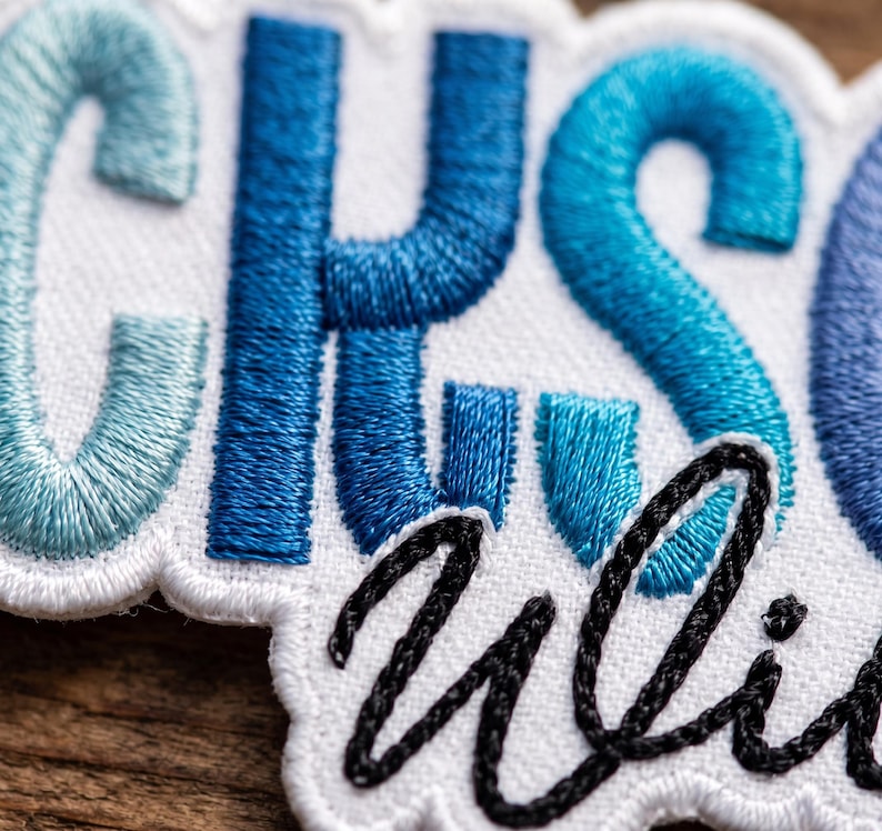 May include: An embroidered patch featuring the letters CHS in varying shades of blue thread on a white background. Below the blue letters, the word uli is stitched in black cursive thread. The patch has a white scalloped border edge.