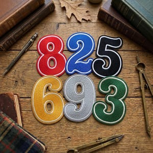 May include: A collection of embroidered number patches in various colors: red, blue, black, yellow, gray, and green. Each number is outlined in white and sits on a wooden surface, surrounded by books, a compass, and a pen.