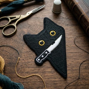 May include: Black cat-shaped embroidered patch with yellow eyes and a white knife. The patch is on a wooden surface with sewing supplies, including scissors, thread, and a thimble. The cat's expression is mischievous.