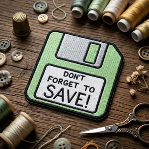 May include: A green and white embroidered patch in the shape of a floppy disk with the text "DON'T FORGET TO SAVE!". The patch is surrounded by sewing supplies, including spools of thread, buttons, and scissors, on a wooden surface.