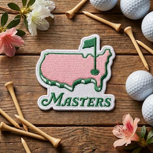 May include: Embroidered patch featuring the Masters logo, a pink outline of a map with a green flag and the word "Masters" in green. The patch is surrounded by golf balls, wooden tees, and flowers on a wooden surface.