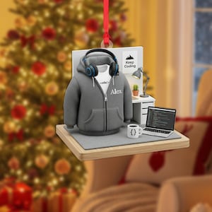 May include: A Christmas ornament featuring a miniature programmer's workspace. It includes a gray hoodie with "Alex" on it, headphones, a laptop displaying code, a coffee cup, and a sign that says "Keep Coding".