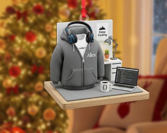 May include: A Christmas ornament featuring a miniature programmer's workspace. It includes a gray hoodie with "Alex" on it, headphones, a laptop displaying code, a coffee cup, and a sign that says "Keep Coding".