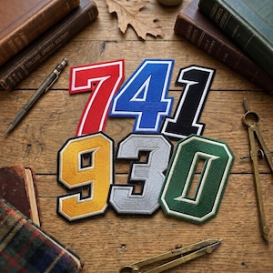May include: A collection of embroidered number patches in various colors, including red, blue, black, yellow, gray, and green. The patches are arranged on a wooden surface with books, a compass, and a pen.