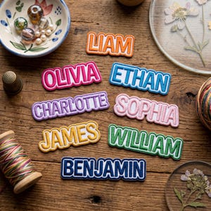 May include: Embroidered name patches in various colors, including orange, pink, blue, purple, yellow, green, and navy. Names include Liam, Olivia, Ethan, Charlotte, Sophia, James, William, and Benjamin. Patches are on a wooden surface.