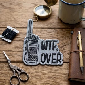 May include: Gray embroidered patch in the shape of a walkie-talkie with the words "WTF OVER" in black. The patch is on a wooden surface, alongside a compass, a mug, a notebook, scissors, and a spool of thread.