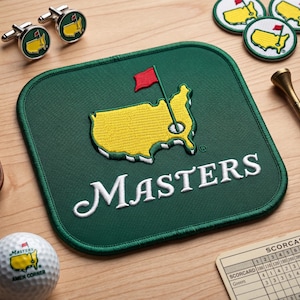 May include: A green square golf course mat with the word "MASTERS" in white script. The mat features a yellow embroidered outline of a golf course with a red flag. Also shown are golf ball, cufflinks, and a scorecard.