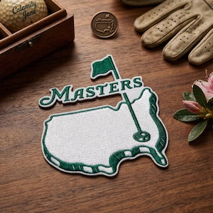 May include: An embroidered patch in the shape of the United States with the word "Masters" in green script. A golf ball, a Masters pin, and a golf glove are also visible. The items are arranged on a wooden surface.