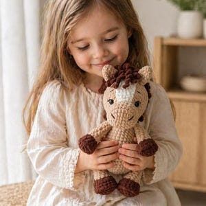 Könnte beinhalten: Gehäkeltes Pferd-Puppe in Beige und Braun. Die Puppe hat einen weißen Streifen im Gesicht und dunkelbraune Hufe und Mähne. Die Puppe wird von einer Person gehalten, die ein cremefarbenes Kleid trägt.