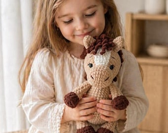 May include: A crocheted horse doll in tan and brown. The doll has a white blaze on its face and dark brown hooves and mane. The doll is being held by a person wearing a cream-colored dress.