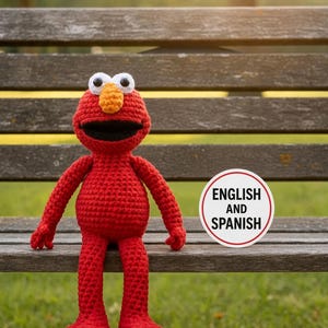 May include: A red crocheted doll with a large orange nose, white eyes with black pupils, and a wide black mouth. Next to the doll is a circular white sign with a red border containing the black text "ENGLISH AND SPANISH" in capital letters.