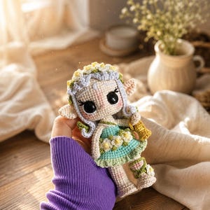 May include: A crochet doll featuring a light-skinned face, large black eyes, and pointed ears. The doll wears a teal dress adorned with white and yellow flowers, a matching floral crown, and light-colored footwear with green accents.