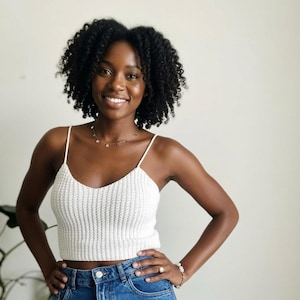 May include: A white ribbed crochet crop top with thin spaghetti straps and a V-neckline, paired with blue denim jeans. The top features a textured knit pattern and a fitted silhouette that ends above the waistline, showcasing a simple design.