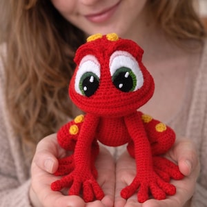 Puede incluir: Una rana roja de ganchillo hecha a mano con grandes ojos con borde blanco e iris verdes. La rana tiene manchas amarillas y se sostiene en manos abiertas. Este juguete caprichoso es un artículo único hecho a mano.