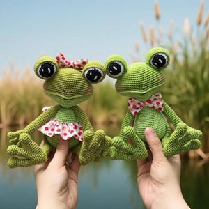 May include: Two handmade green crochet frog dolls. One frog wears a red and white polka dot bow and the other wears a matching bow tie and a red and white polka dot dress. The frogs have large black eyes and are posed in front of a body of water.