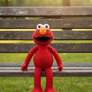 Könnte beinhalten: Handgemachte, rote Häkelpuppe mit lächelndem Gesicht, weißen Augen und einer orangenen Nase. Die Puppe sitzt auf einer Holzbank in einem Park. Die Arme und Beine der Puppe sind ausgestreckt.