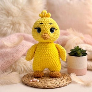 Puede incluir: Una muñeca de pollito de ganchillo amarillo hecha a mano con un pequeño mechón de pelo, ojos azules y un pico marrón. El pollito está de pie sobre una estera tejida. La muñeca es un juguete suave y lindo.