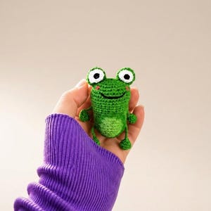 Op de afbeelding: Een klein, handgemaakt gehaakt kikkerfiguurtje met een felgroen lichaam, een lichtgroene buikvlek en grote witte ogen met zwarte pupillen. De kikker heeft een geborduurde zwarte glimlach, roze wangetjes en kleine armen en benen.