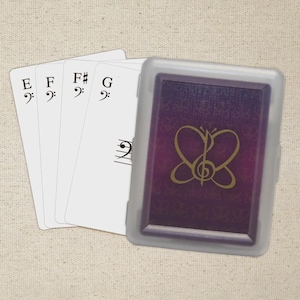 May include: A deck of playing cards with musical notes and a butterfly design. The cards are white with black musical symbols and letters E, F, F#, and G. The card case is purple with a gold butterfly design.