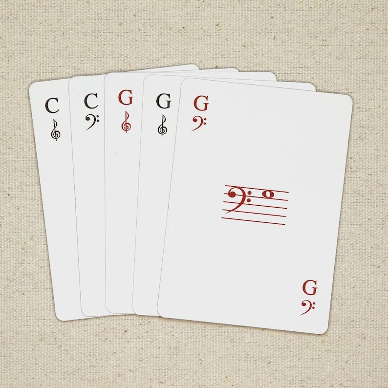 May include: A set of five white playing cards with musical notation. The cards feature treble and bass clef symbols, along with the letters "C" and "G" in red. The cards are arranged on a neutral-toned fabric surface.