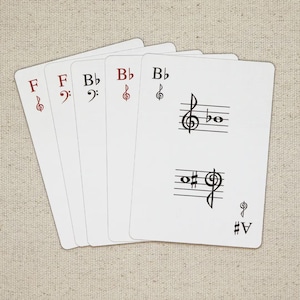 May include: Five white playing cards with musical notation. The cards display musical symbols such as treble and bass clefs, flats, and sharps. The cards are arranged in a fan shape, with the top card showing a treble clef and a musical staff.