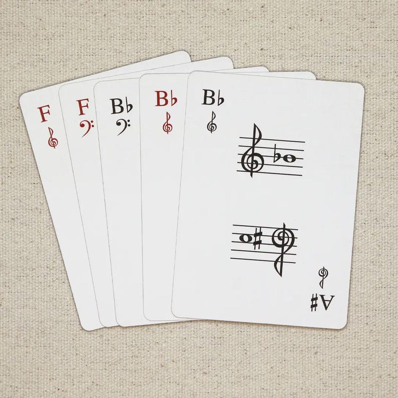 May include: Five white playing cards with musical notation. The cards display musical symbols such as treble and bass clefs, flats, and sharps. The cards are arranged in a fan shape, with the top card showing a treble clef and a musical staff.