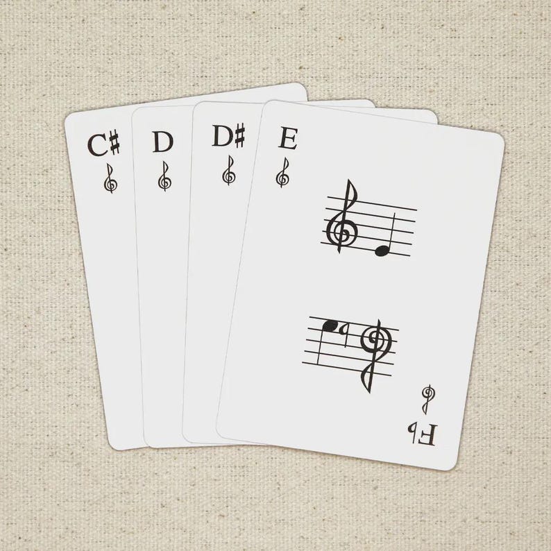 May include: Four white playing cards with musical notes and symbols. The cards display the notes C#, D, D#, and E, along with treble clef symbols. One card features musical notation. The background is a beige fabric.