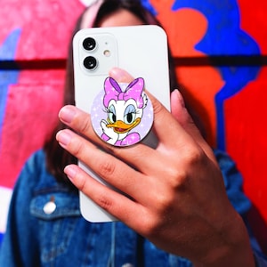 May include: A round phone grip featuring Daisy Duck from Disney. The grip has a light purple background with sparkling stars. Daisy Duck is wearing a pink bow and dress. The phone grip is attached to a white smartphone.