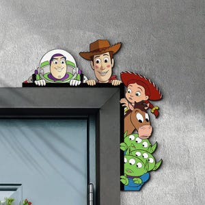 Puede incluir: Recortes de madera de los personajes de Toy Story Buzz Lightyear, Woody, Jessie, los Aliens y Bullseye. Los personajes se asoman por encima de un marco de puerta negro, con una pared gris de fondo. Los recortes son coloridos y detallados.