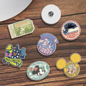 May include: A selection of enamel pins with Disney characters and travel themes. The pins feature Mickey Mouse, Donald Duck, and references to California, Florida, and Michigan. The pins are in various shapes and sizes, with bright colours.