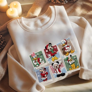 May include: White sweatshirt featuring embroidered patches of Disney characters in festive holiday attire. Mickey Mouse, Donald Duck, Daisy Duck, Goofy, and Pluto are depicted in Christmas-themed designs. The sweatshirt has a crew neck and long sleeves.