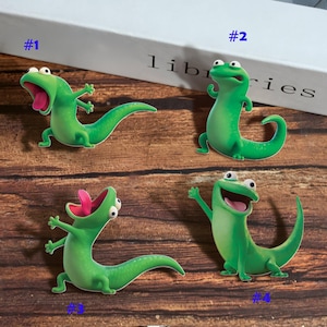 May include: Four green cartoon lizard figurines with white eyes and open mouths. The lizards have long tails and are posed in different positions. The figurines are on a wooden surface.