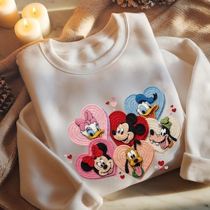 May include: White sweatshirt featuring embroidered heart-shaped patches with Disney characters Mickey Mouse, Minnie Mouse, Donald Duck, Daisy Duck, Goofy, and Pluto. The patches are in various colors, including red, pink, blue, and yellow.