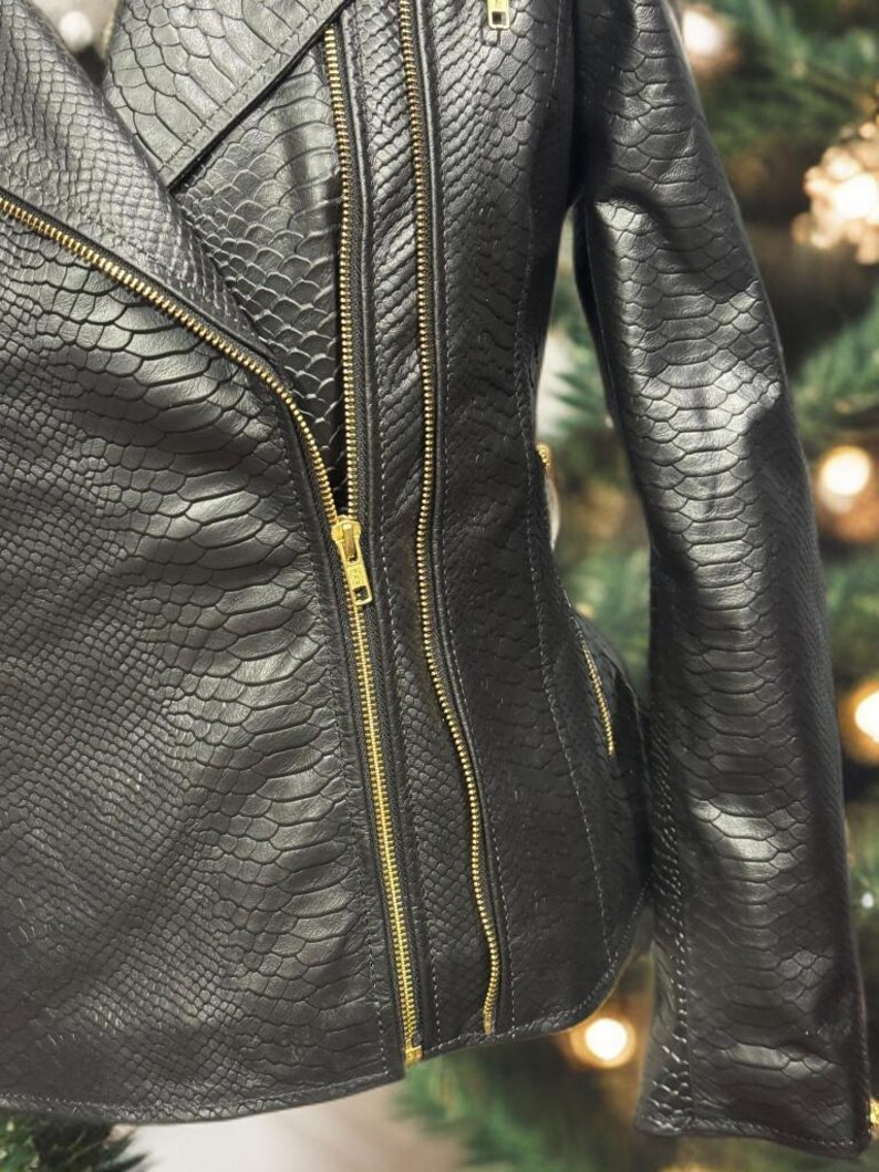 K&ouml;nnte beinhalten: Schwarze Kunstlederjacke mit Schlangenlederstruktur. Die Jacke hat mehrere goldfarbene Rei&szlig;verschl&uuml;sse und ein klassisches Bikerjacken-Design. Die Nahaufnahme zeigt die Details des Materials und der Hardware.