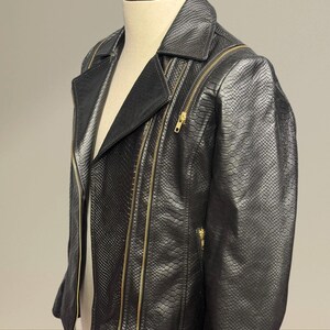K&ouml;nnte beinhalten: Schwarze Kunstlederjacke mit Schlangenlederstruktur. Die Jacke hat einen klassischen Biker-Stil mit Reverskragen, mehreren Rei&szlig;verschlusstaschen und goldfarbenen Rei&szlig;verschl&uuml;ssen. Die Jacke wird auf einer Schaufensterpuppe pr&auml;sentiert.