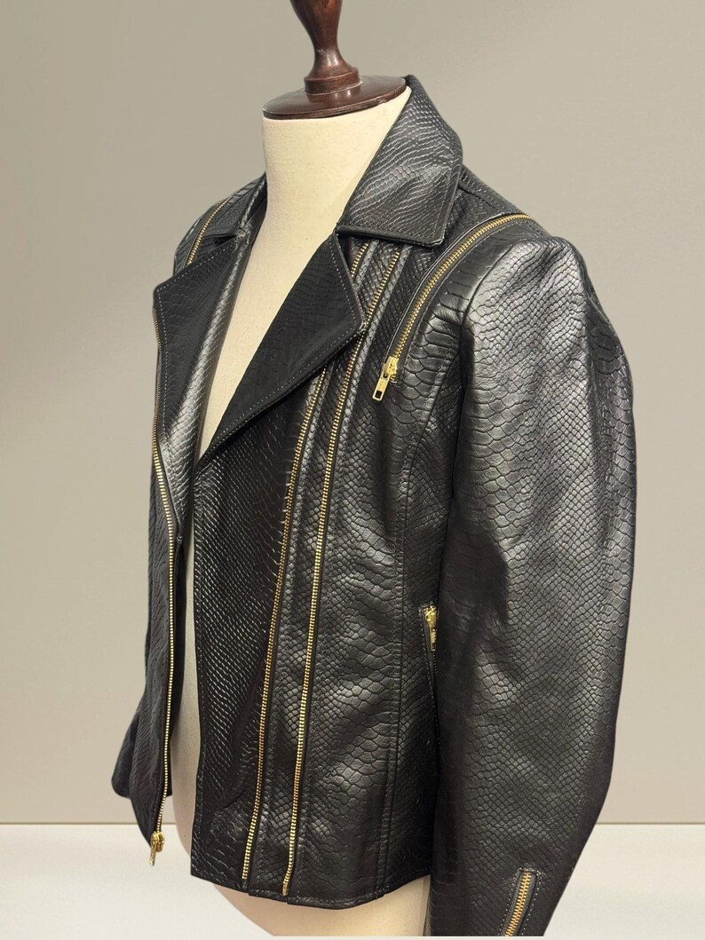 K&ouml;nnte beinhalten: Schwarze Kunstlederjacke mit Schlangenlederstruktur. Die Jacke hat einen klassischen Biker-Stil mit Reverskragen, mehreren Rei&szlig;verschlusstaschen und goldfarbenen Rei&szlig;verschl&uuml;ssen. Die Jacke wird auf einer Schaufensterpuppe pr&auml;sentiert.