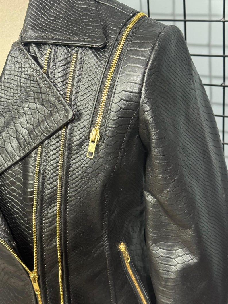 May include: Black faux leather jacket with a snakeskin texture. The jacket features a classic asymmetrical design with a notched lapel, multiple zippered pockets, and gold-toned zippers. The jacket is a stylish outerwear piece.