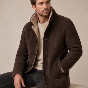 May include: A dark brown jacket with a high collar and a light beige interior lining. The jacket features a button-front closure with four visible dark buttons and two side pockets. It is worn over a light beige knit sweater and dark grey jeans.