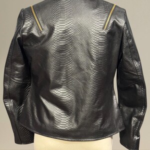 K&ouml;nnte beinhalten: Schwarze Kunstlederjacke mit Schlangenlederstruktur. Die Jacke hat einen Reverskragen, lange &Auml;rmel und goldfarbene Rei&szlig;verschl&uuml;sse an den Schultern. Die Jacke wird auf einer Schaufensterpuppe pr&auml;sentiert.