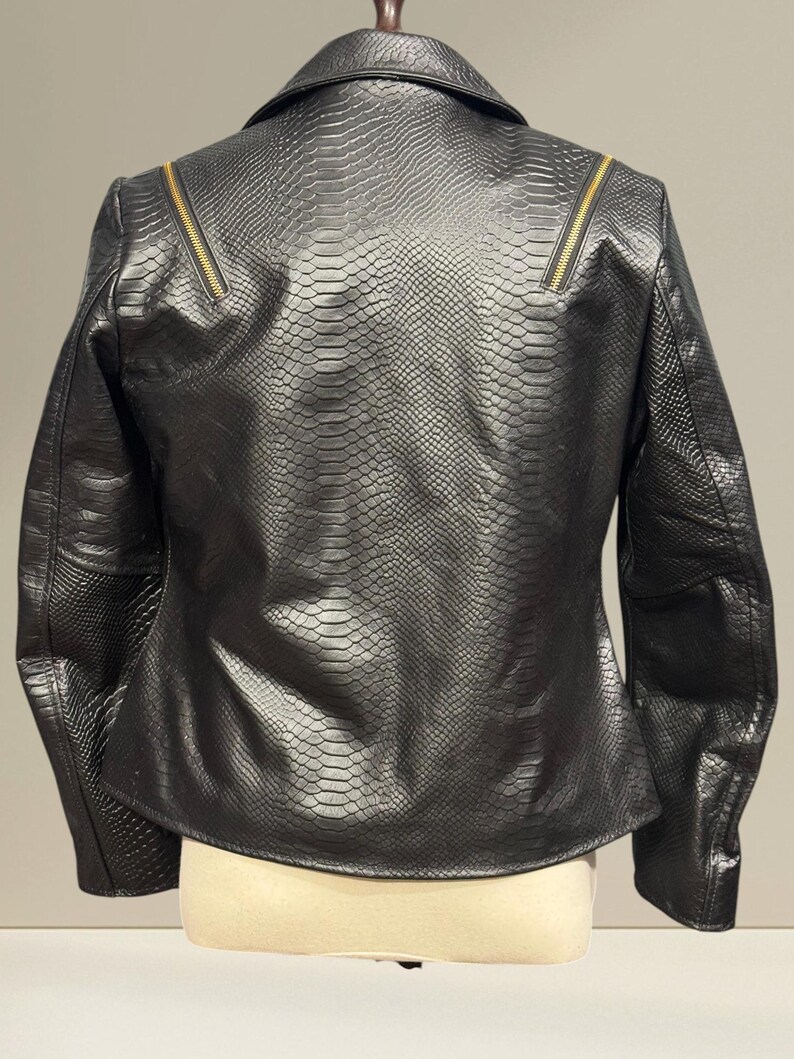 K&ouml;nnte beinhalten: Schwarze Kunstlederjacke mit Schlangenlederstruktur. Die Jacke hat einen Reverskragen, lange &Auml;rmel und goldfarbene Rei&szlig;verschl&uuml;sse an den Schultern. Die Jacke wird auf einer Schaufensterpuppe pr&auml;sentiert.