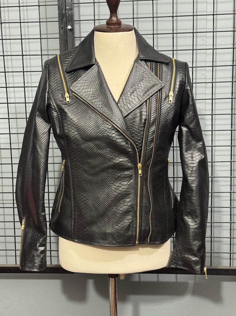 May include: Black faux leather jacket with a snakeskin texture and gold zippers. The jacket has a classic asymmetrical design with a notched collar and multiple zippered pockets. The jacket is displayed on a mannequin.