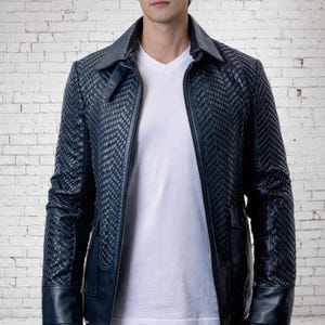 May include: A dark blue jacket featuring a textured, woven chevron pattern on the front and sleeves, a solid collar, and smooth cuffs. The jacket is worn over a plain white V-neck t-shirt and dark trousers, showcasing a structured design.