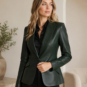 May include: A dark forest green leather blazer featuring a single-button closure, notched lapels, and two front flap pockets. The jacket is worn over a black collared shirt and black trousers, showcasing a structured fit with long sleeves.