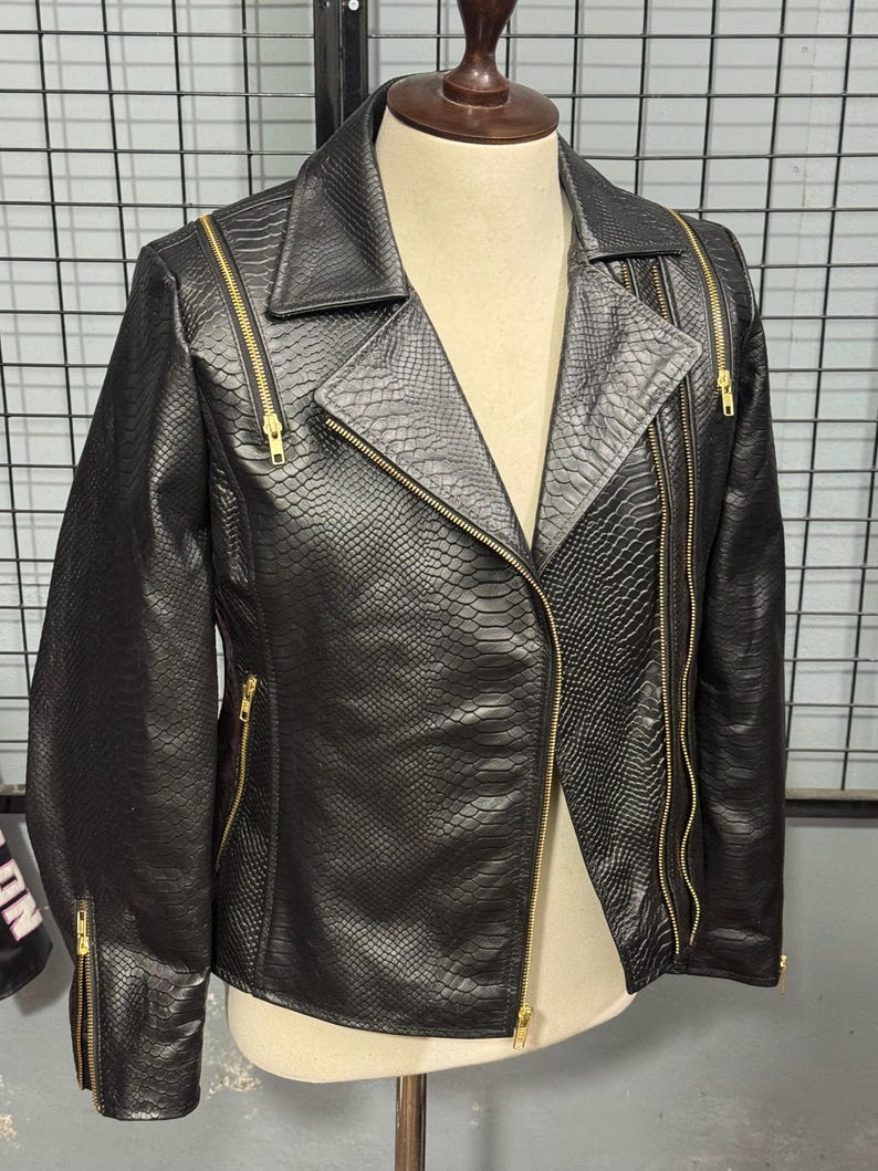 May include: Black faux leather jacket with a snakeskin texture. The jacket features a classic asymmetrical zip closure, notched lapels, and multiple zippered pockets. Gold-toned zippers and hardware provide a striking contrast to the dark material. The jacket is displayed on a mannequin.