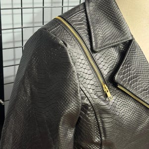 May include: Black faux leather jacket with a snakeskin texture. The jacket features a classic asymmetrical zipper design with gold-colored hardware. The collar is a notched lapel style, and the jacket is displayed on a mannequin.