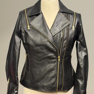 K&ouml;nnte beinhalten: Schwarze Lederjacke mit Schlangenmuster und goldenen Rei&szlig;verschl&uuml;ssen. Die Jacke hat einen klassischen Biker-Stil mit asymmetrischem Rei&szlig;verschluss, Reverskragen und mehreren Rei&szlig;verschlusstaschen. Die Jacke wird auf einer Schaufensterpuppe pr&auml;sentiert.