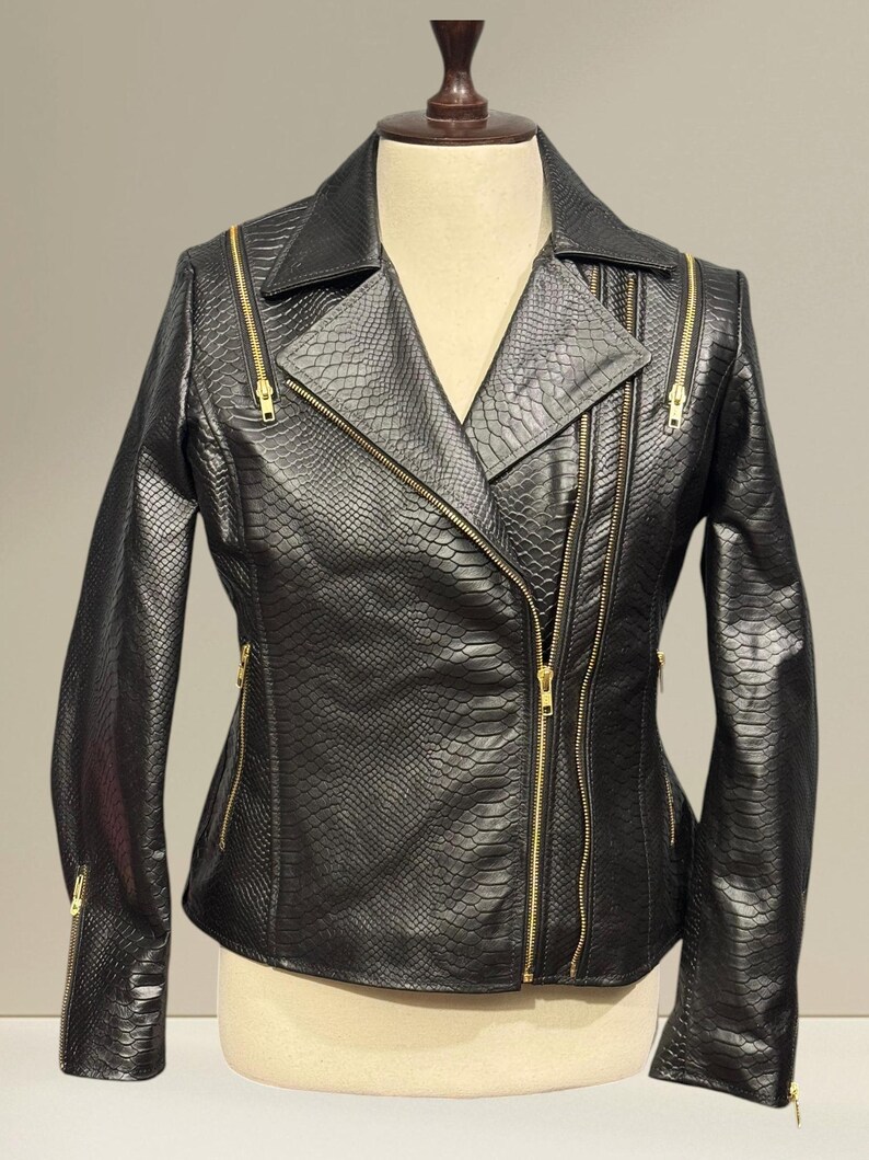 K&ouml;nnte beinhalten: Schwarze Lederjacke mit Schlangenmuster und goldenen Rei&szlig;verschl&uuml;ssen. Die Jacke hat einen klassischen Biker-Stil mit asymmetrischem Rei&szlig;verschluss, Reverskragen und mehreren Rei&szlig;verschlusstaschen. Die Jacke wird auf einer Schaufensterpuppe pr&auml;sentiert.