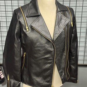 May include: Black faux leather jacket with a snakeskin texture. The jacket features a classic asymmetrical zip closure, notched lapels, and multiple zippered pockets. Gold-toned zippers and hardware provide a striking contrast to the dark material. The jacket is displayed on a mannequin.
