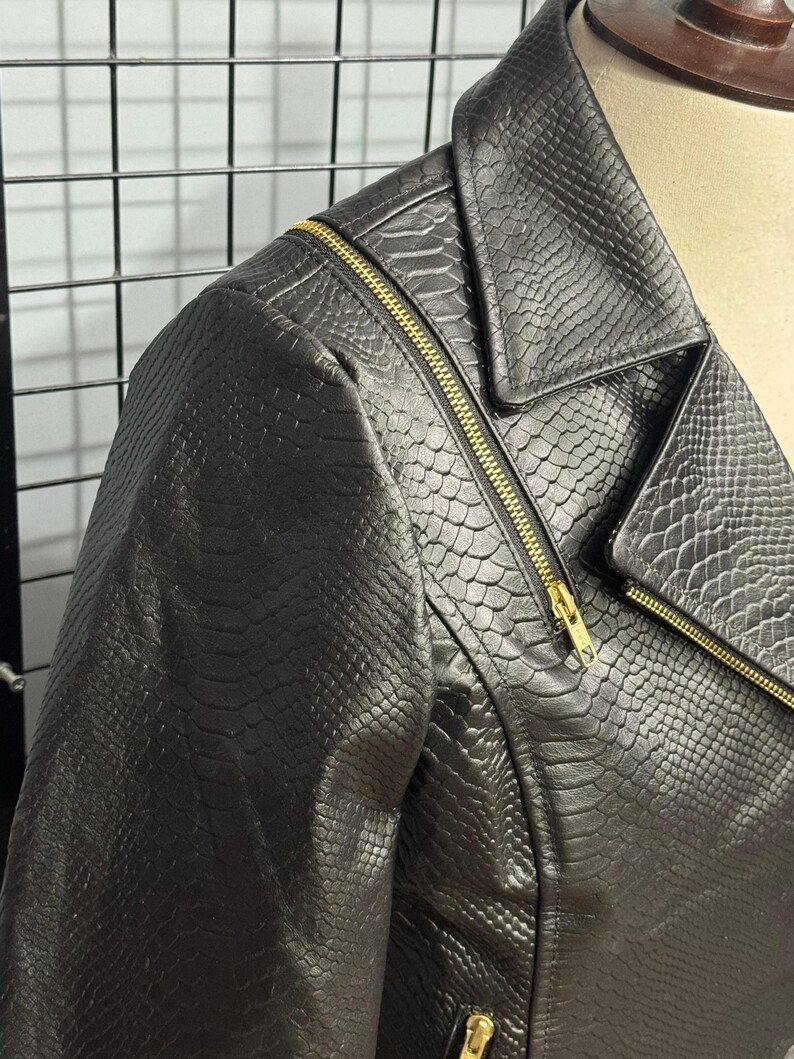 May include: Black faux leather jacket with a snakeskin texture. The jacket features a classic asymmetrical zipper design with gold-colored hardware. The collar is a notched lapel style, and the jacket is displayed on a mannequin.