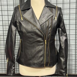 May include: Black faux leather jacket with a snakeskin texture and gold zippers. The jacket has a classic asymmetrical design with a notched collar and multiple zippered pockets. The jacket is displayed on a mannequin.